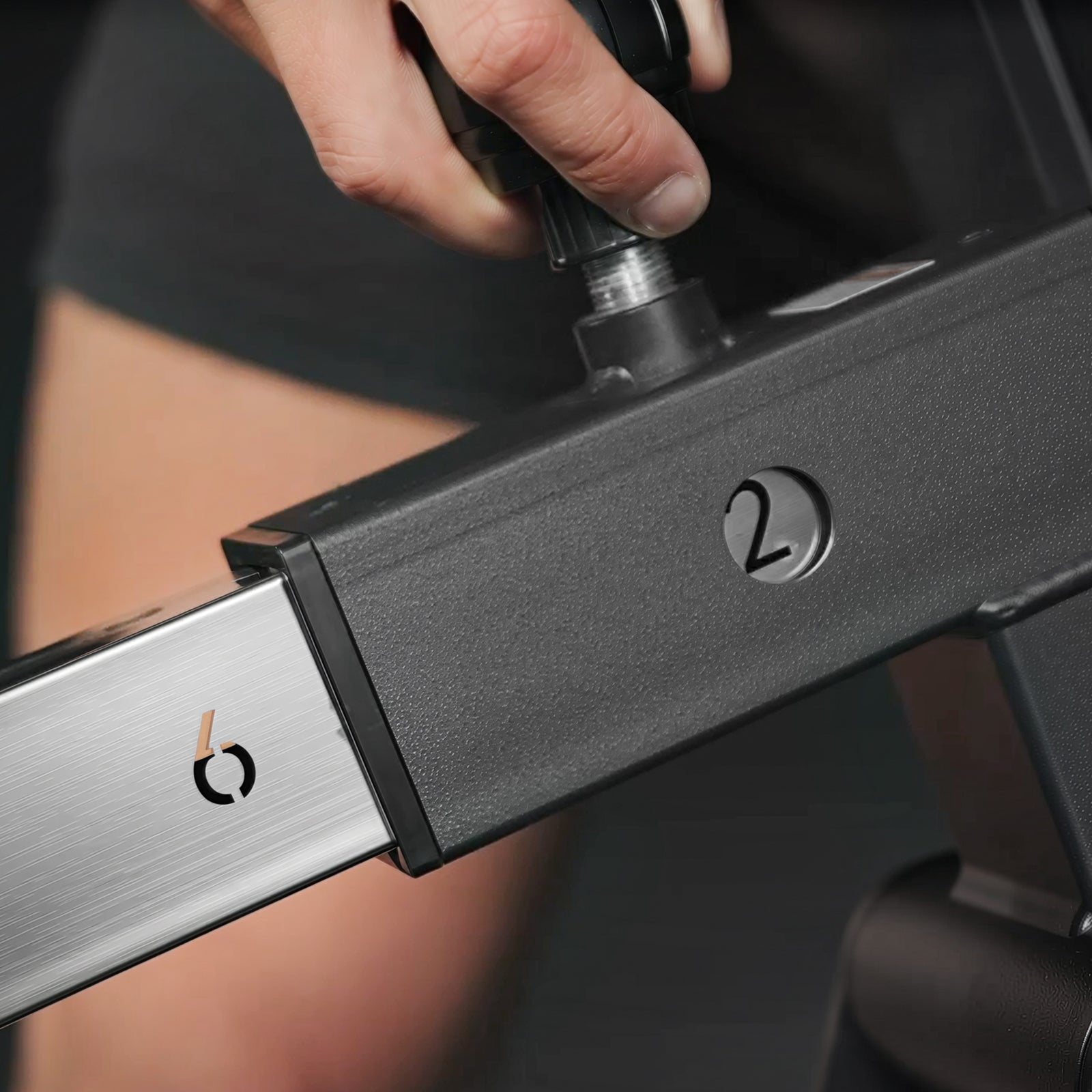 Hand turning a black knob on a textured gym machine frame near a silver sliding bar with a logo and the number 2