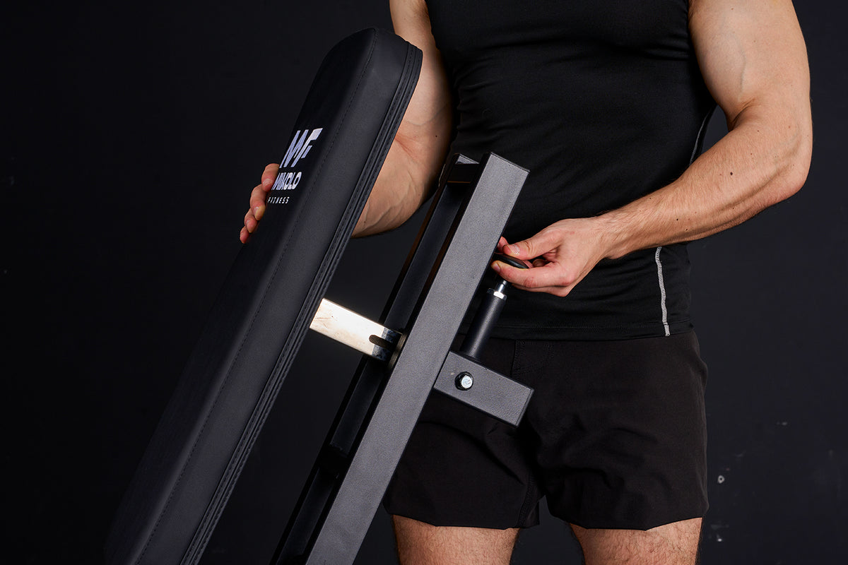 Person in black gym wear holding a black padded leg extension machine part with Mikolo Fitness logo and grey metal frame