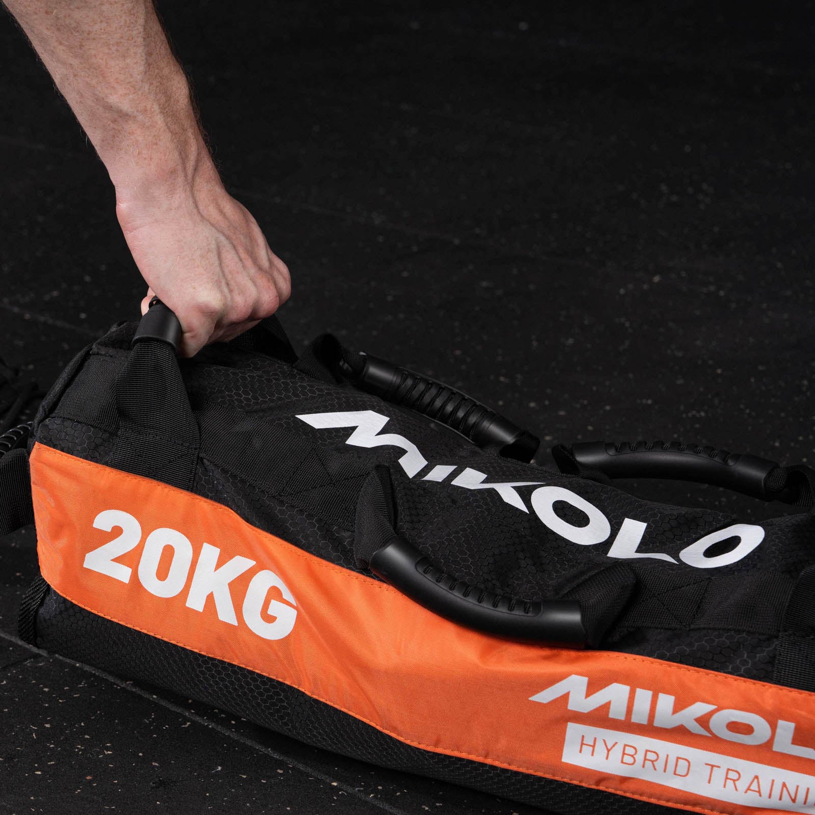 A hand grips the handle of a black and orange Mikolo 20kg sandbag with multiple handles on a gym floor