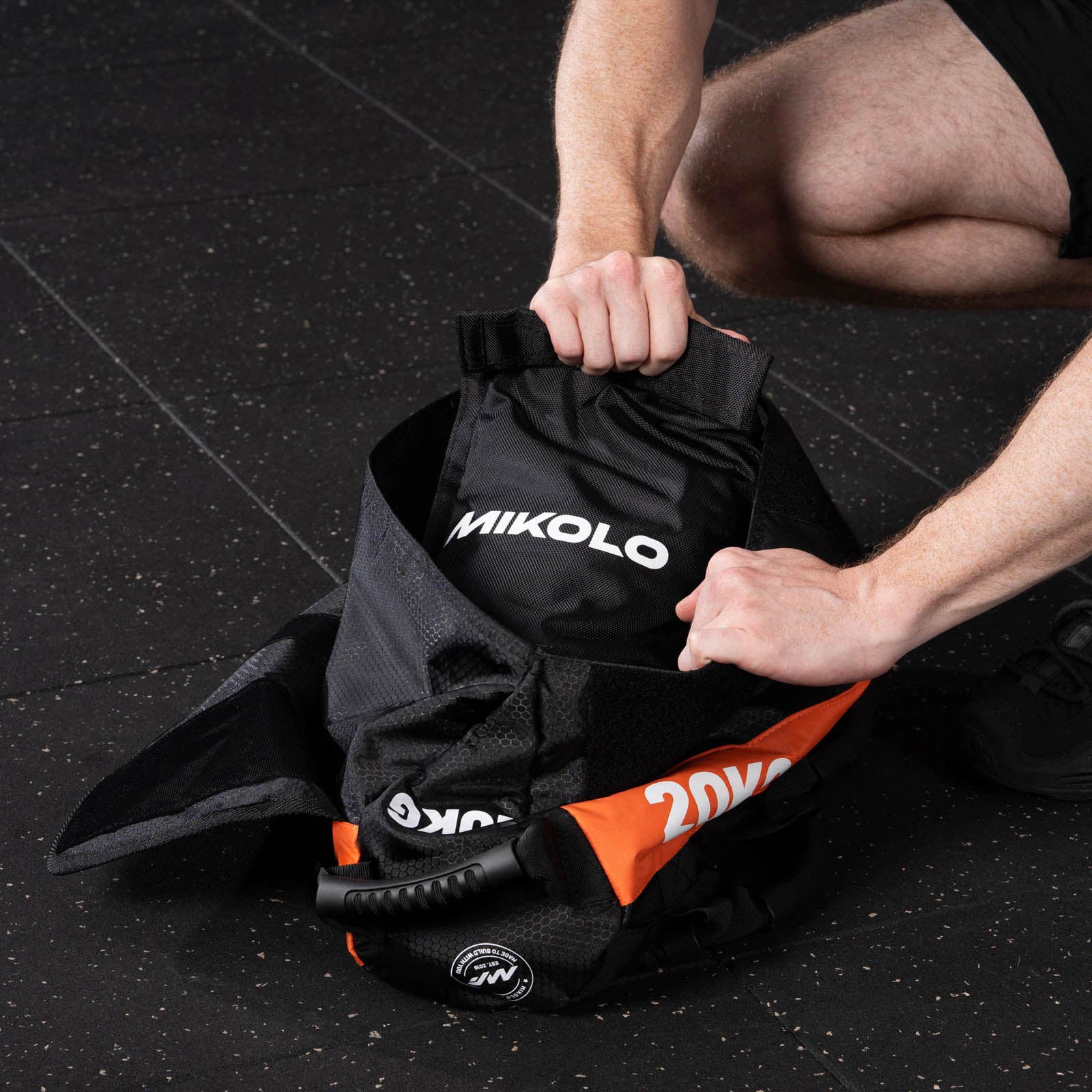 Person placing a black MIKOLO inner bag into an open 20kg orange and black sandbag on a gym floor