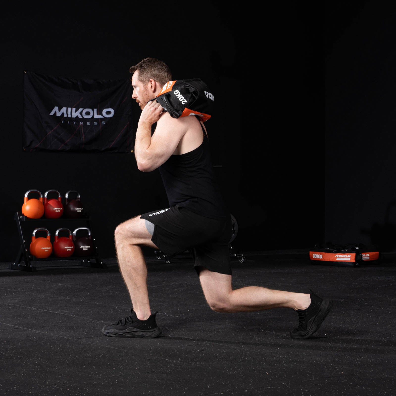 A man performs a lunge carrying a black and orange sandbag on his shoulder in a gym with kettlebells