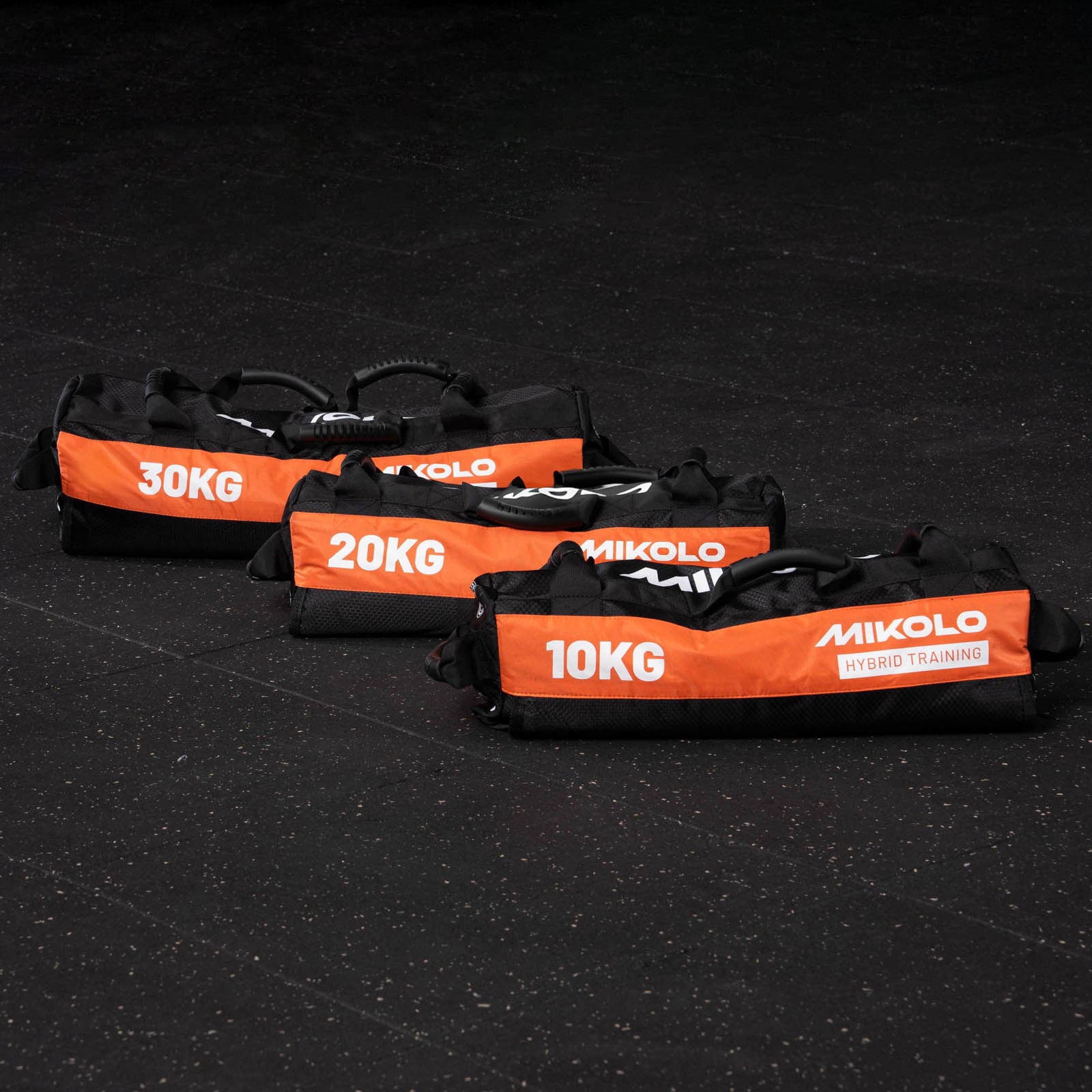 Three black and orange Mikolo sandbags labeled 30kg, 20kg, and 10kg arranged on a dark gym floor