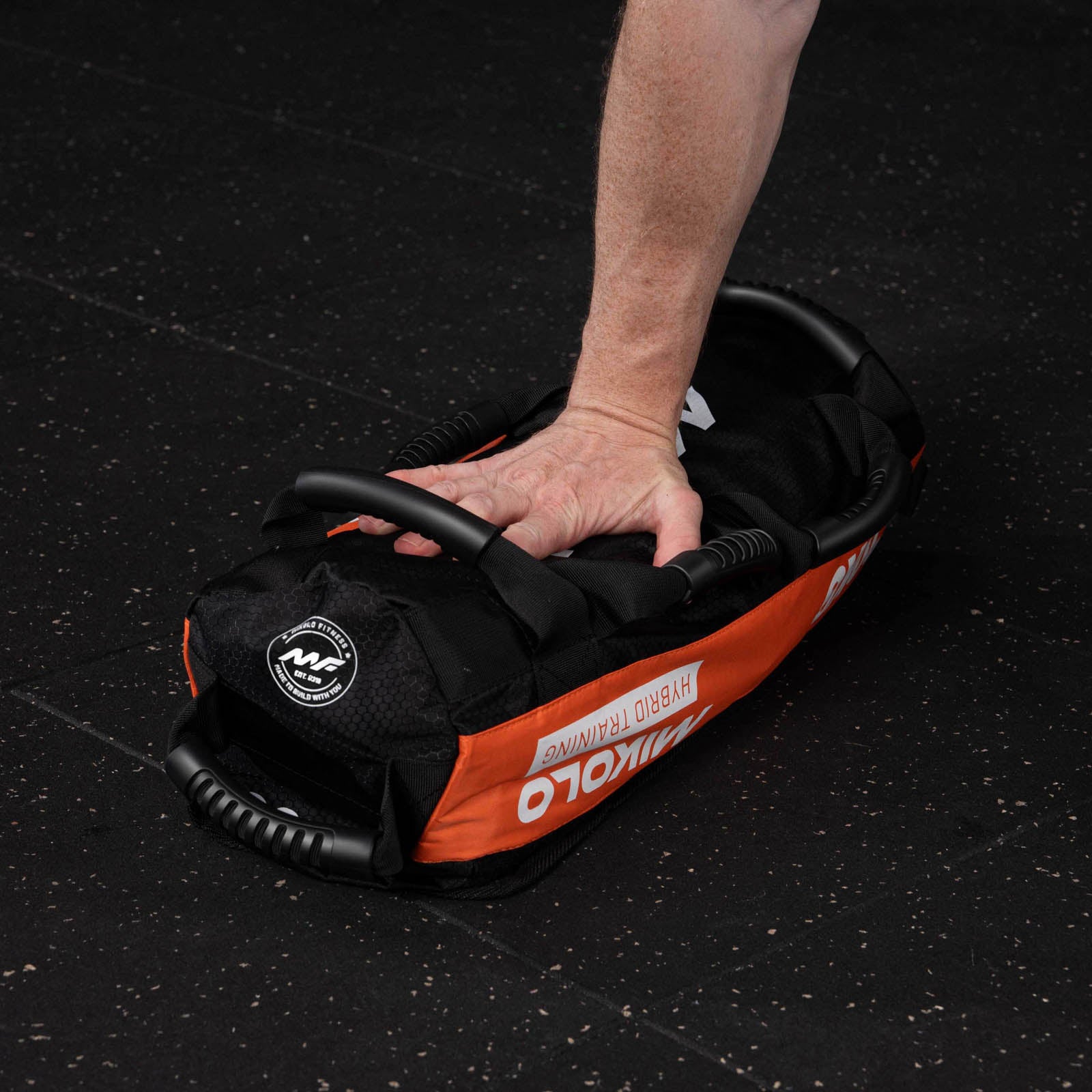 A person's hand gripping the handle of a black and orange Mikolo sandbag resting on a black gym floor