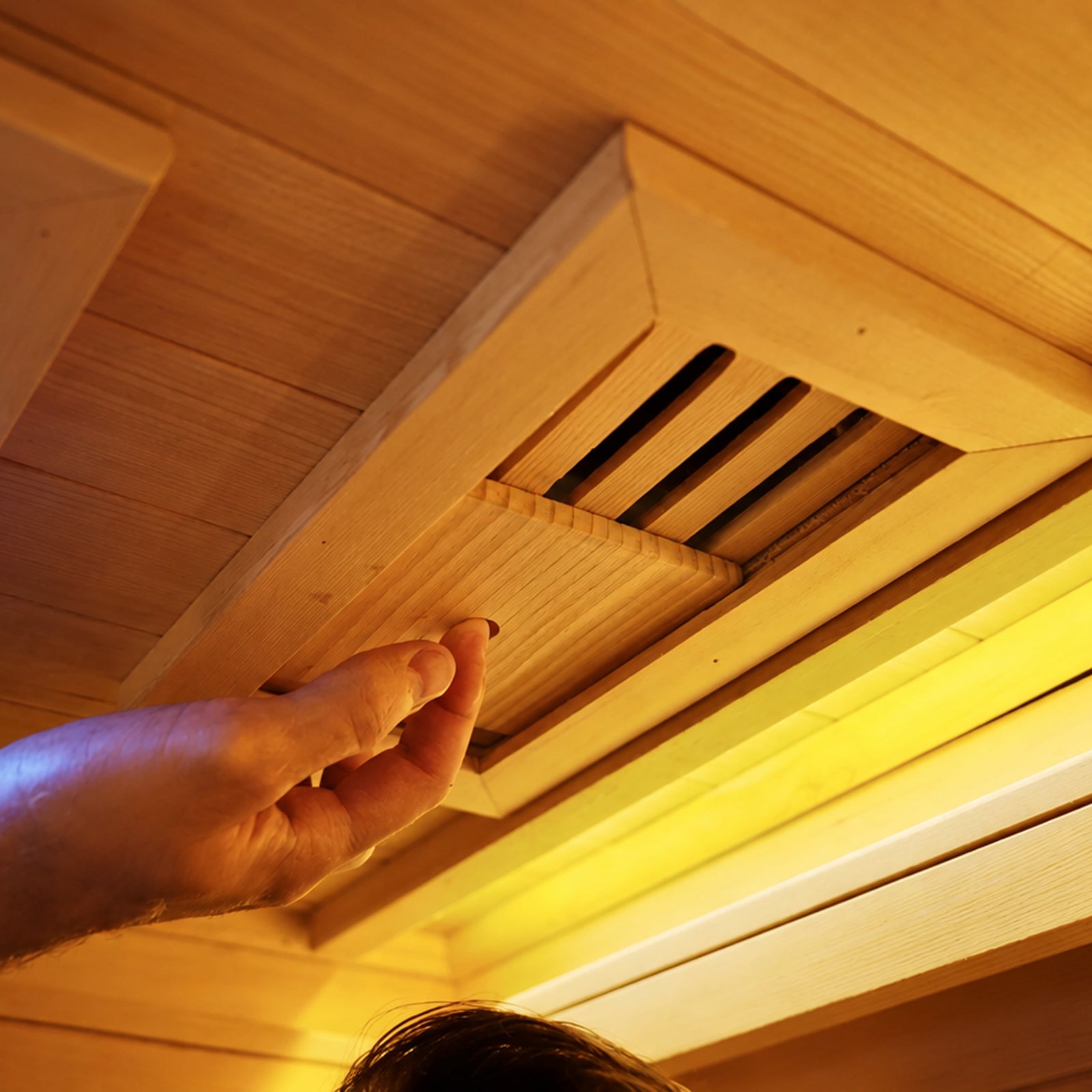 A hand adjusts a wooden sliding vent cover on the ceiling of a sauna near a warm light fixture