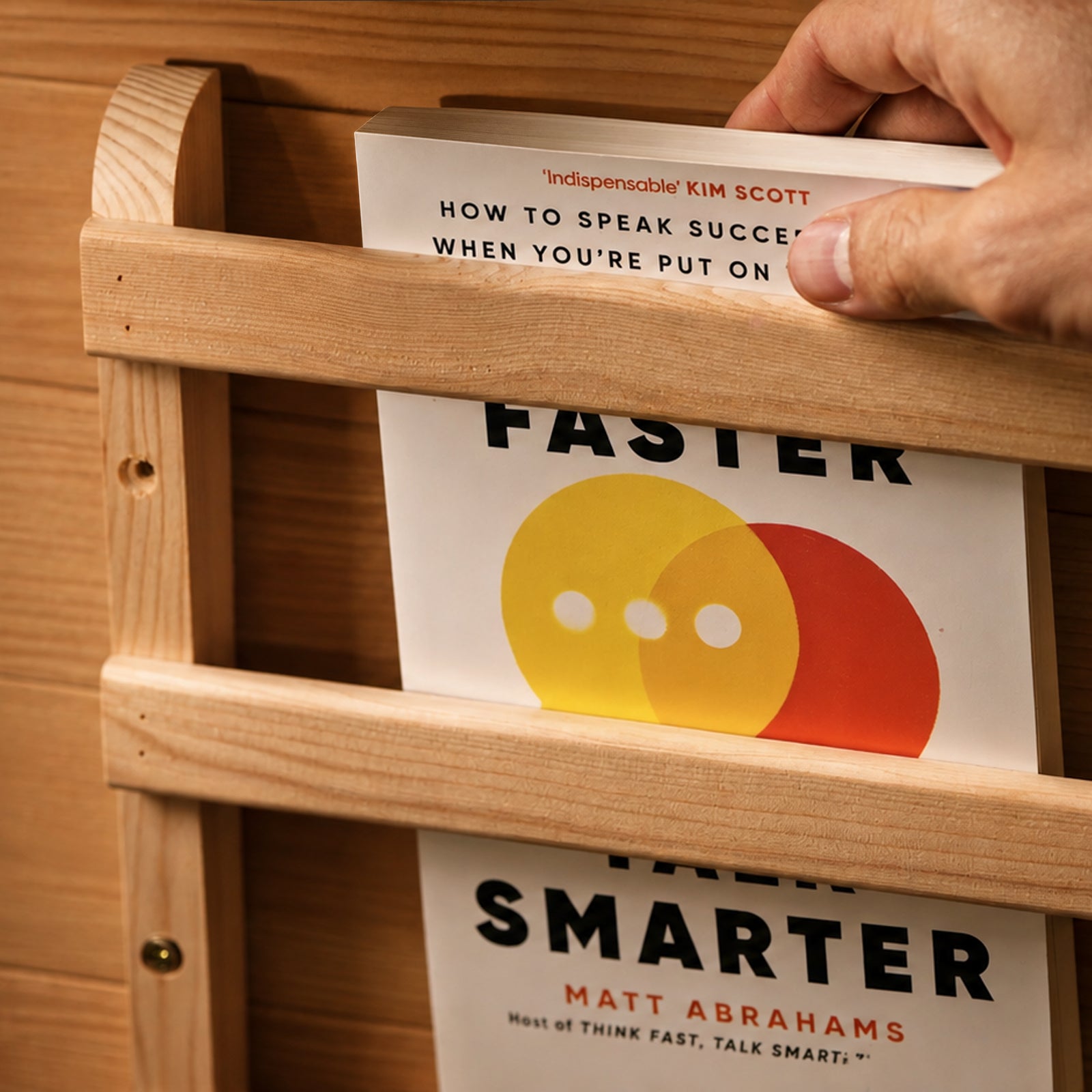 A hand places a book titled Think Faster Talk Smarter by Matt Abrahams into a light wooden wall rack