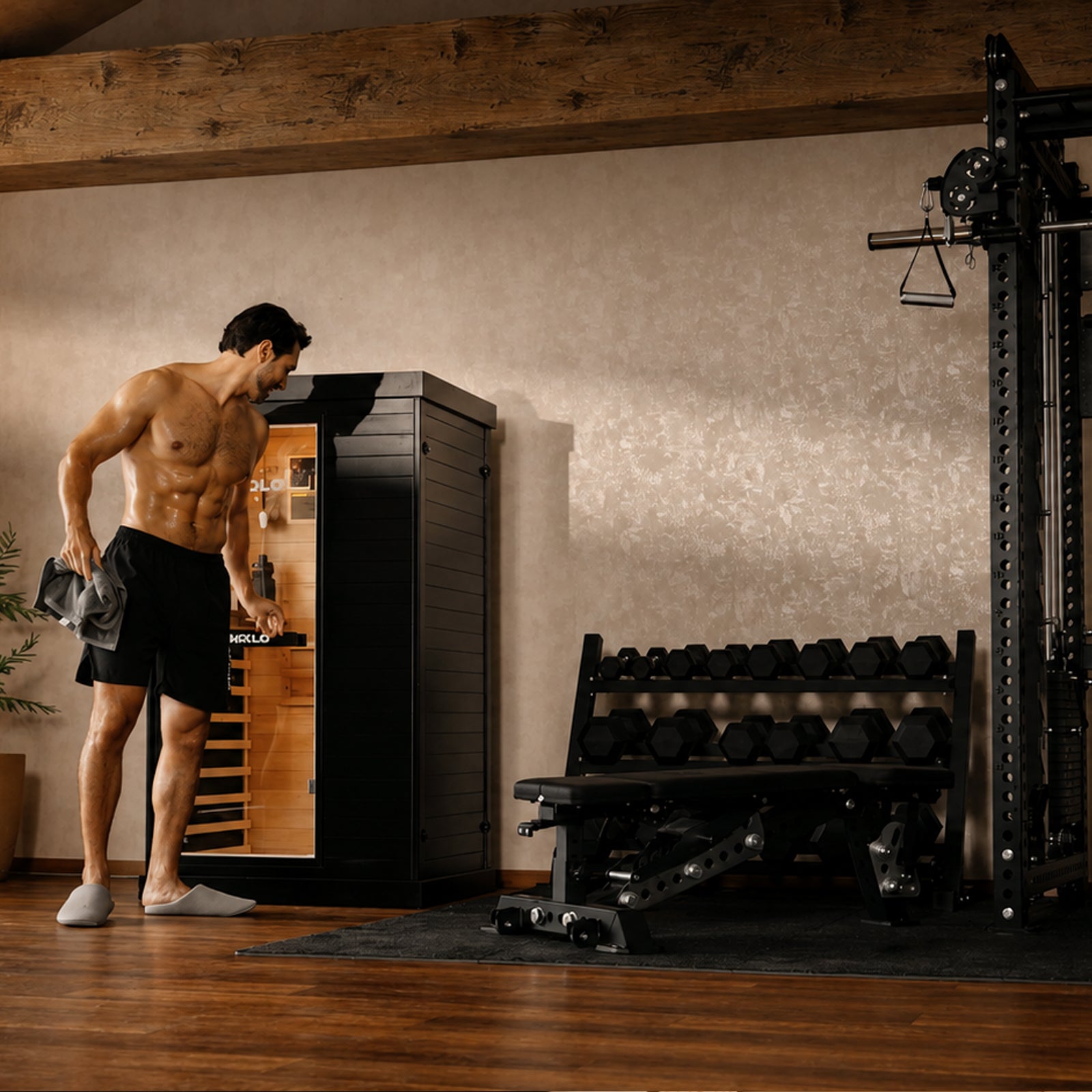 Shirtless man holding a towel stands next to an open black infrared sauna in a home gym with a dumbbell rack and bench