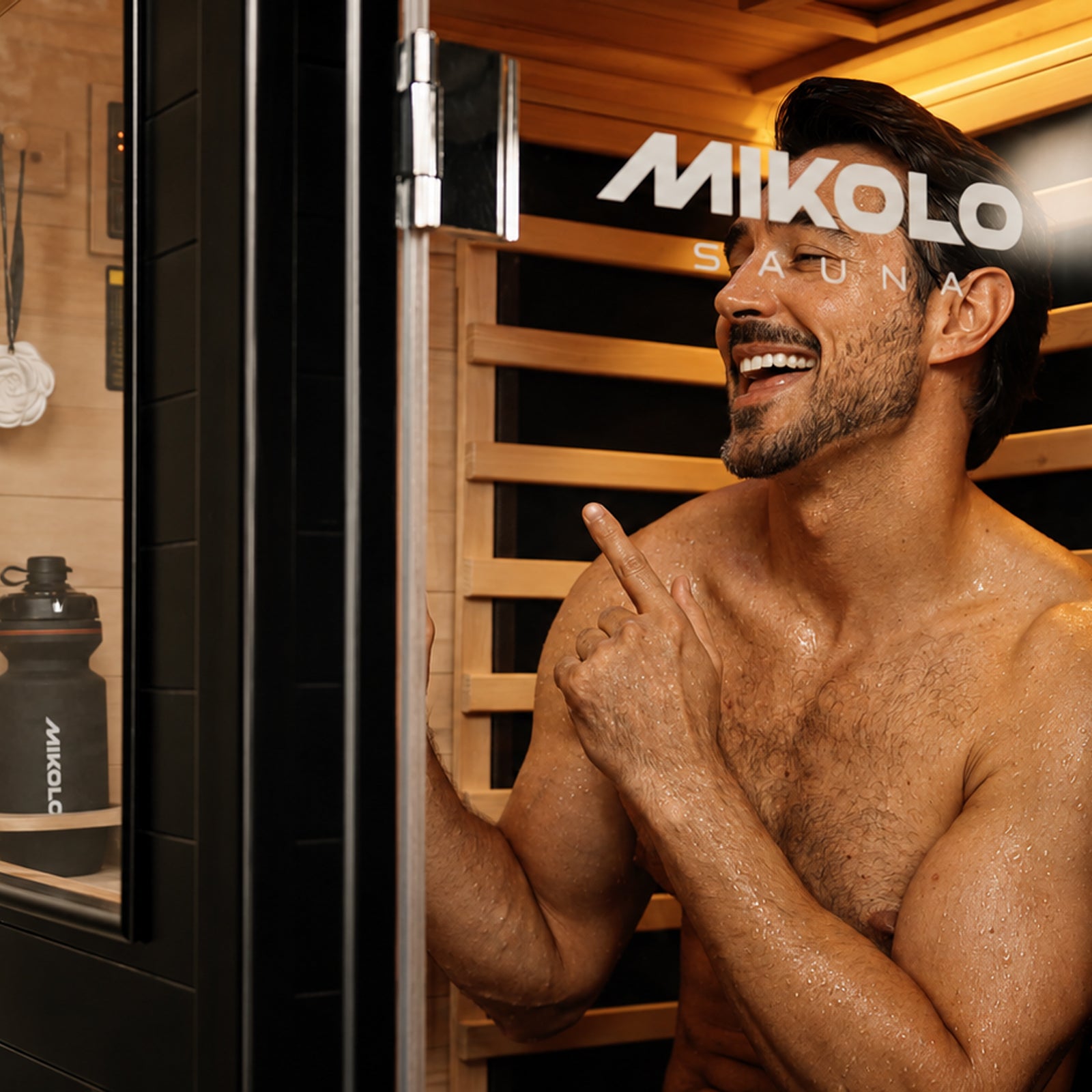 Smiling shirtless man inside a wooden Mikolo sauna pointing, with a branded water bottle on a shelf outside the glass door