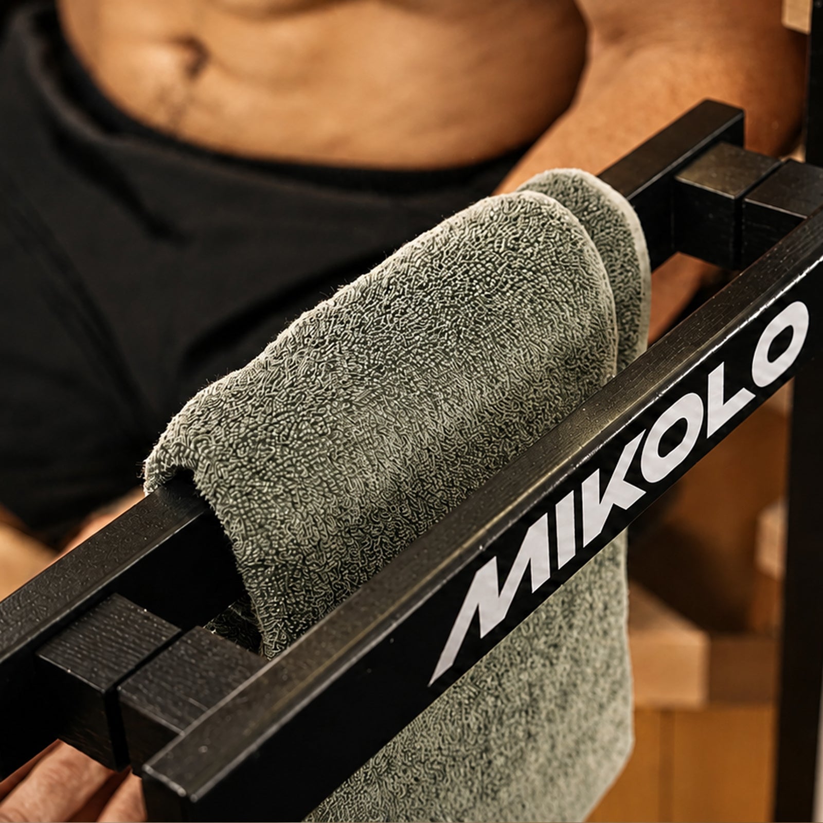 Grey towel draped over a black metal bar featuring the white MIKOLO logo, with a shirtless person's torso in the background