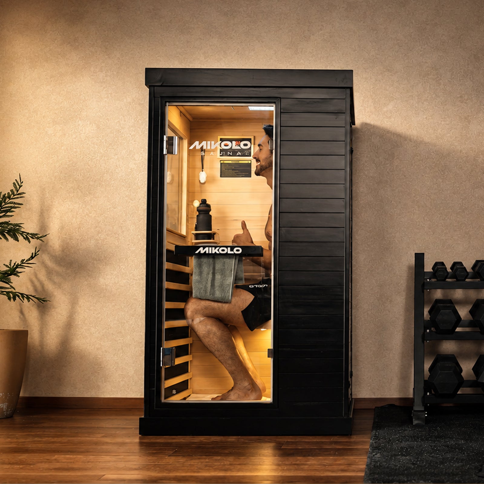 Man sitting inside a black Mikolo infrared sauna giving a thumbs up, with a towel and water bottle, next to dumbbells