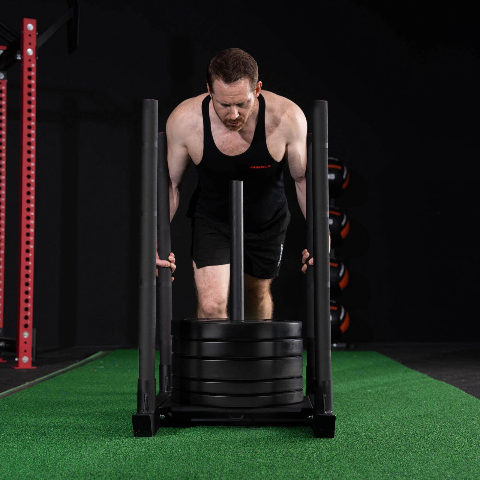Man in black tank top pushing a loaded black weighted sled across green gym turf