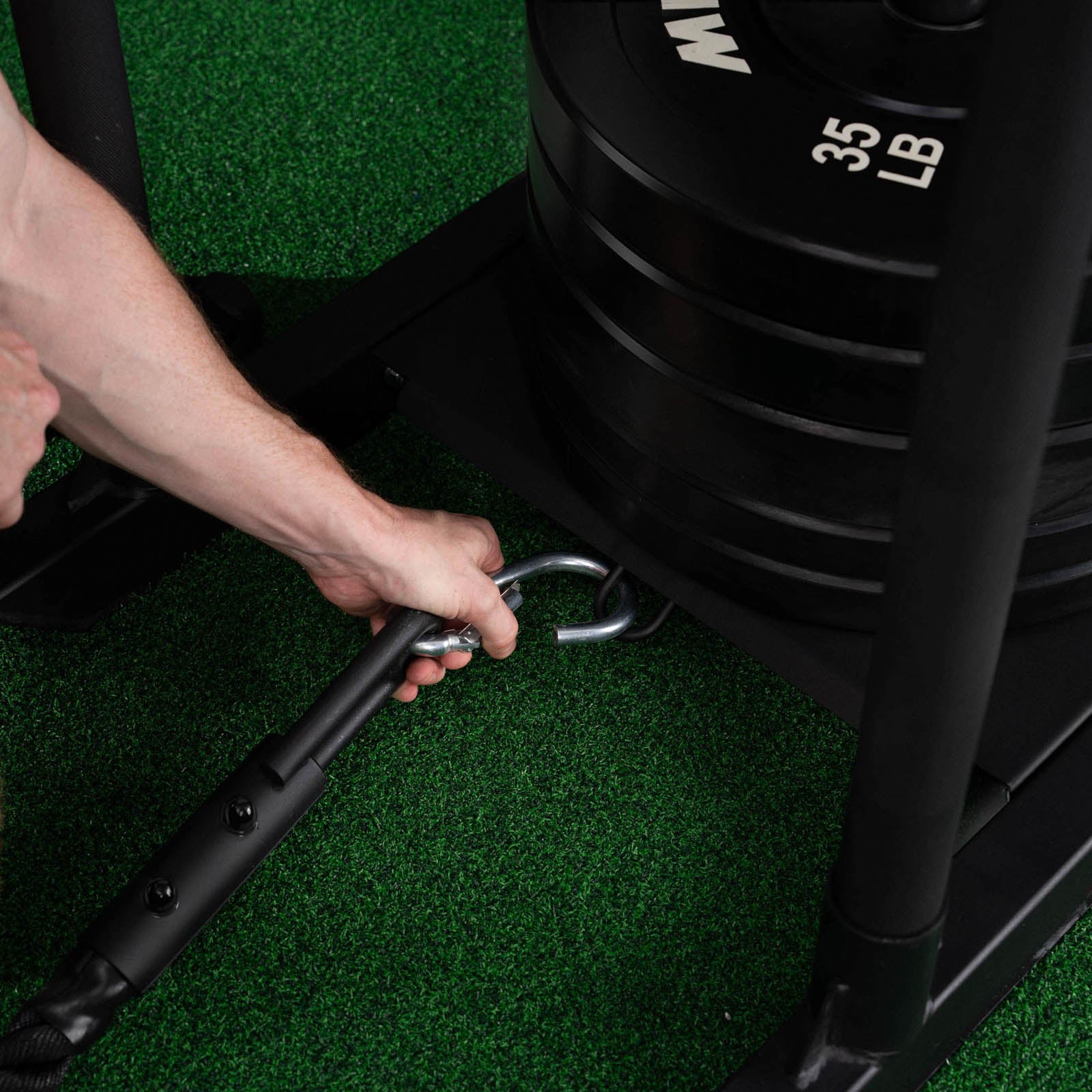 Hands attaching a metal clip from a black rope handle to the frame of a weighted sled with 35 lb plates on green turf