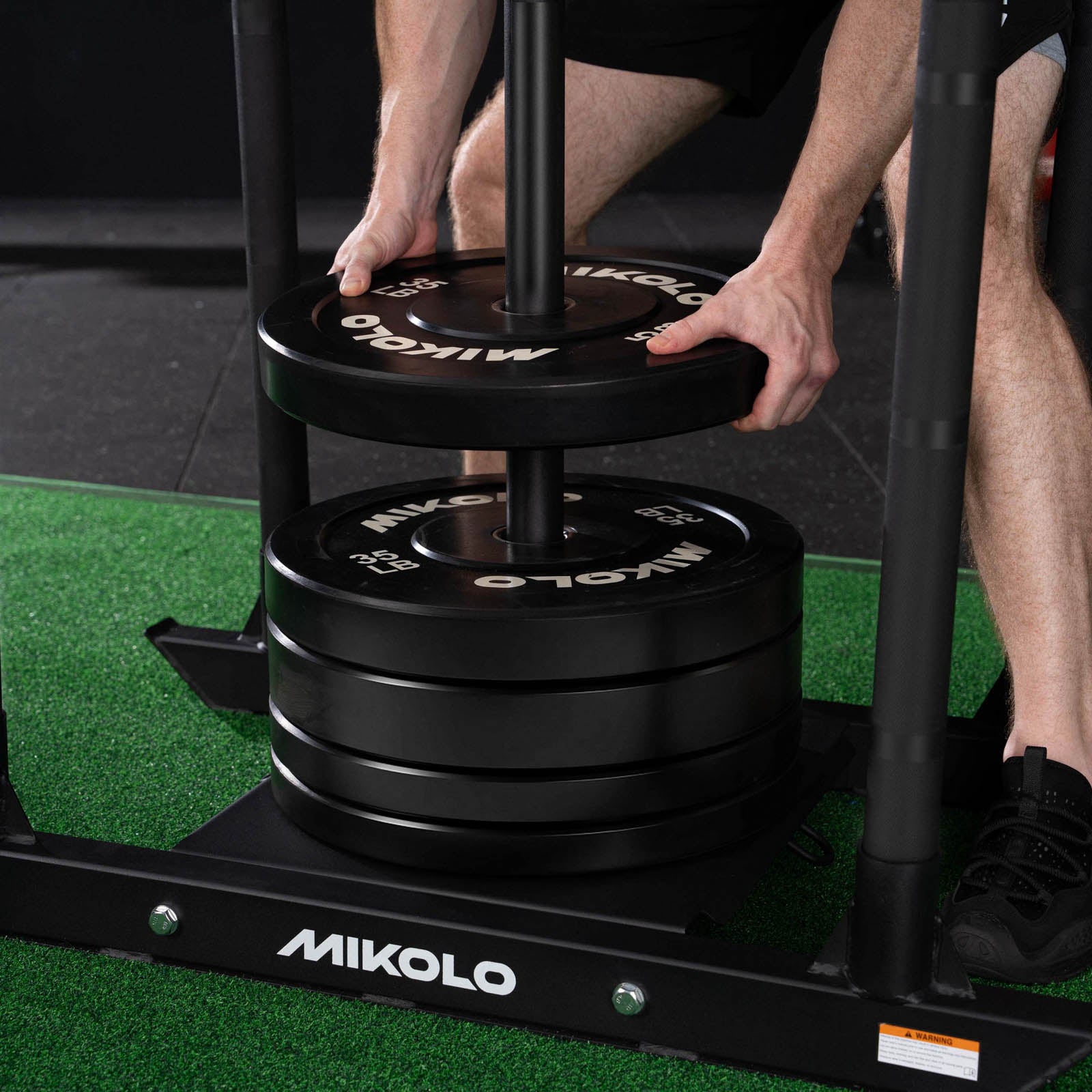 A person loads a black 33 lb Mikolo weight plate onto a sled pole above a stack of weights on green turf
