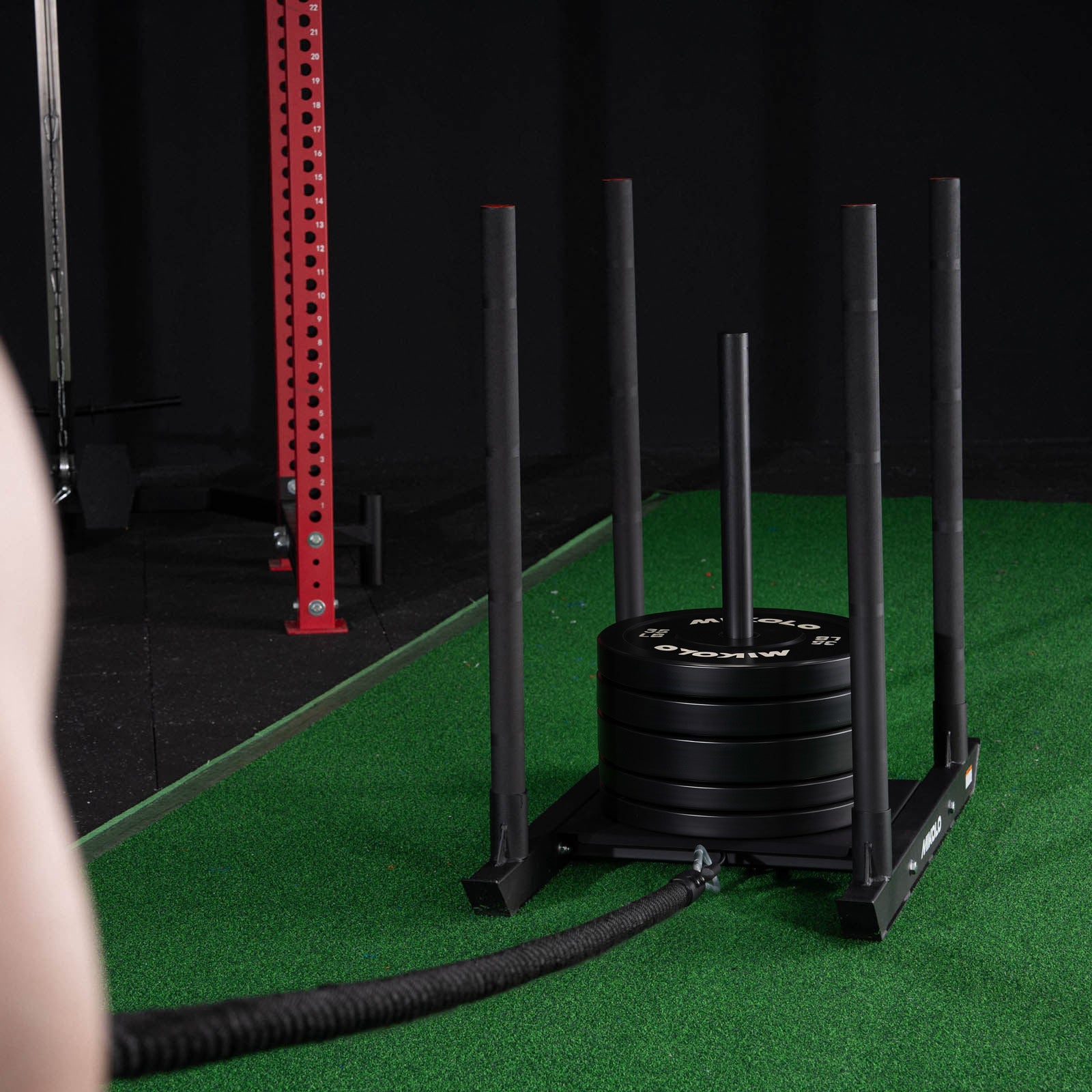 Black Mikolo sled loaded with weight plates on green turf, featuring vertical poles and a pulling rope