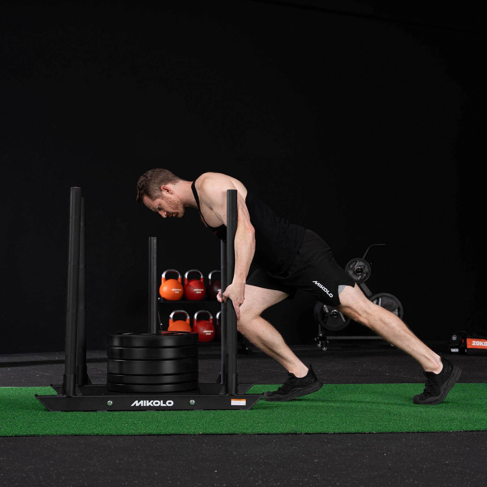 A man pushes a black Mikolo sled loaded with weight plates across green turf