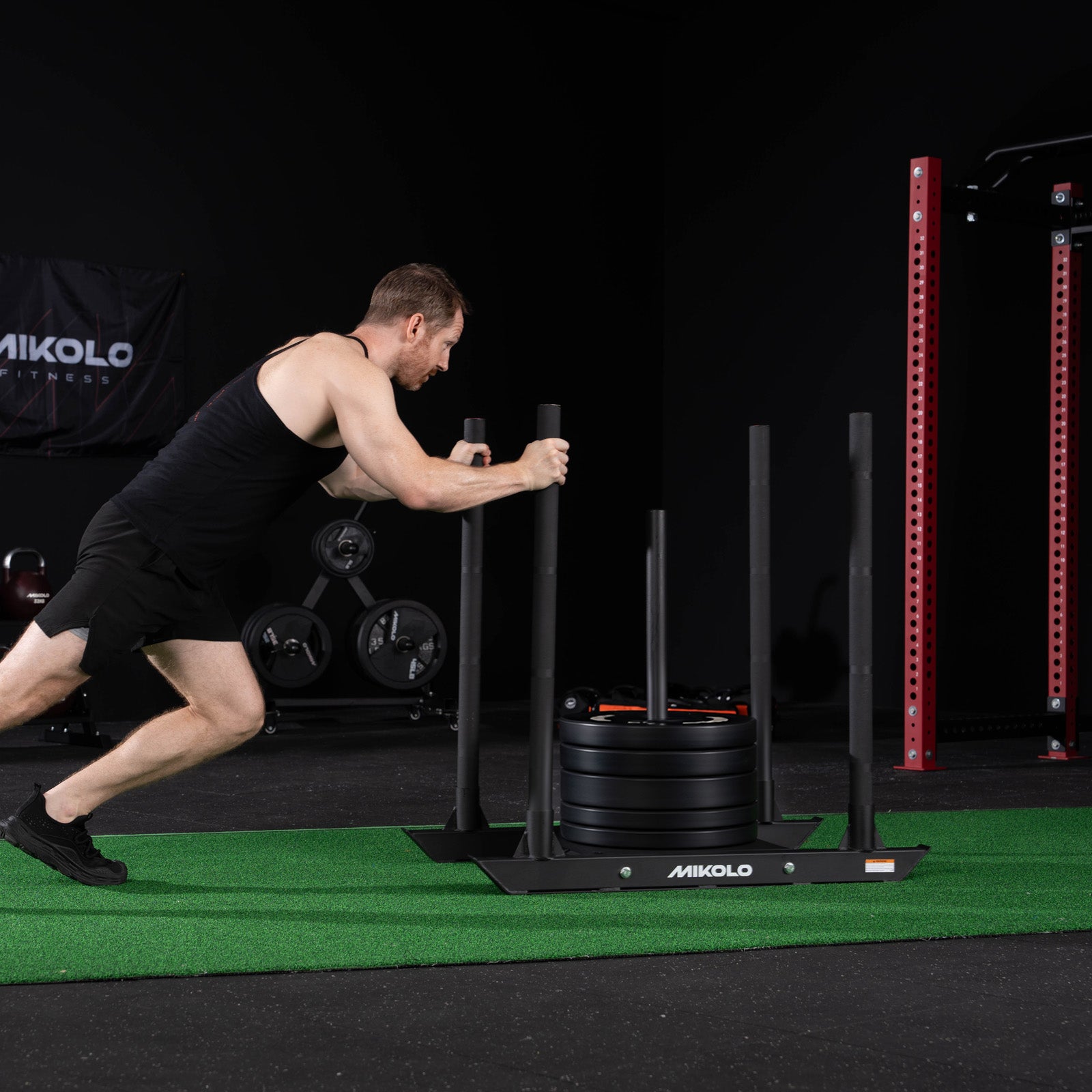 A man in a black tank top pushes a black Mikolo weighted sled loaded with plates across green turf