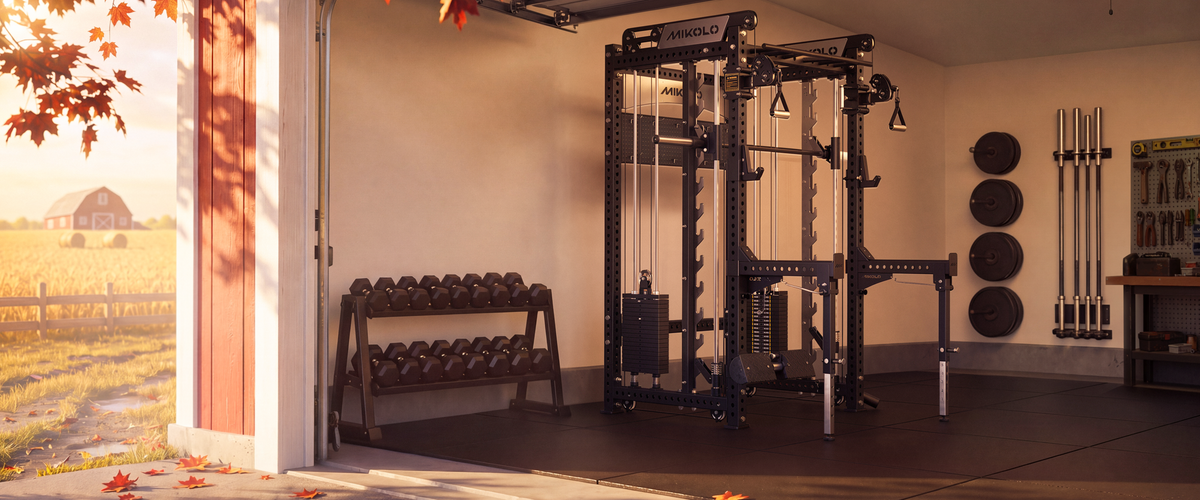 A black and dark gray home gym power rack with a cable pulley system, shown inside a garage with an open door revealing an outdoor autumn scene. A wooden dumbbell rack with multiple dumbbells sits to the left, and weight plates and barbell racks are mounted on the wall to the right.