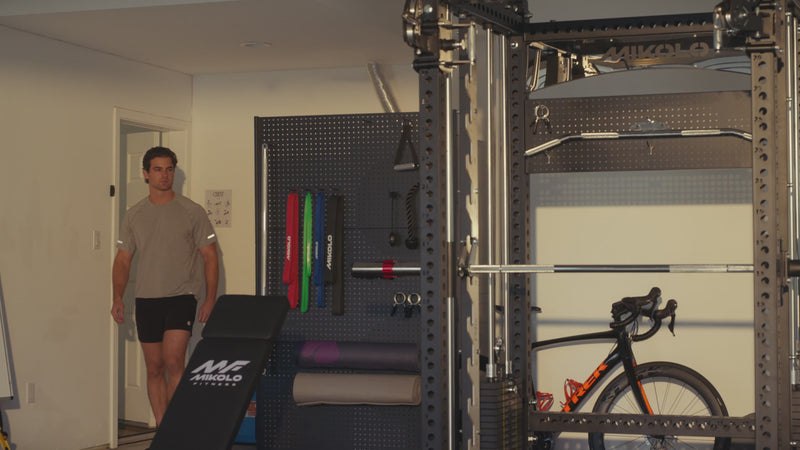 Man entering a home gym with a black MIKOLO power rack, weight bench, pegboard wall, and a Trek bicycle