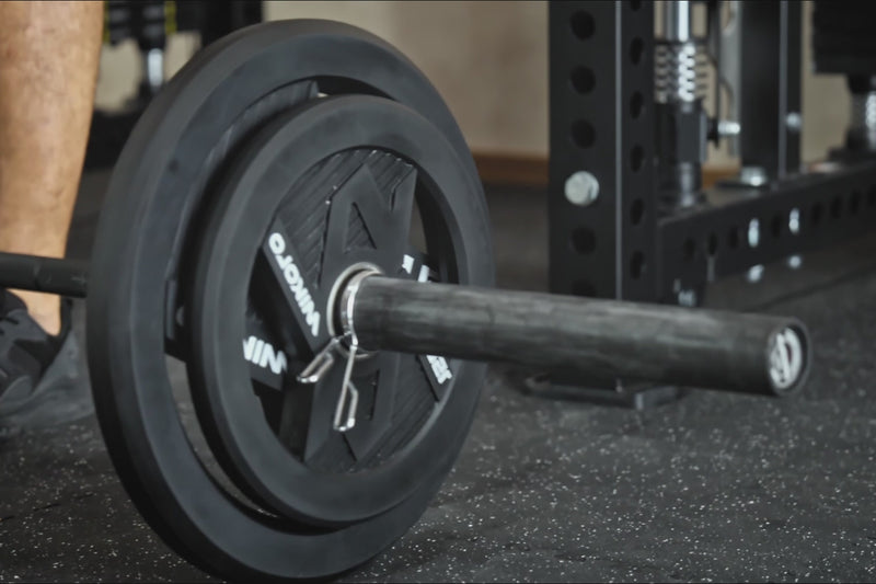 Close-up of a barbell loaded with black bumper plates secured by a spring clip on a rubber gym floor